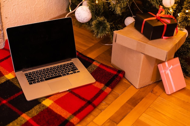 Foto online-shopping zu weihnachten. laptop mit kopienraum auf dem bildschirm lag, geschenkboxen und weihnachtsbaum im hintergrund. internet-handel auf winterferienkonzept