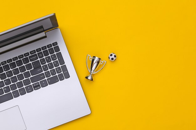 Online-sportwetten. laptop, fußball und meisterpokal auf gelbem hintergrund mit kopienraum. ansicht von oben. flach legen