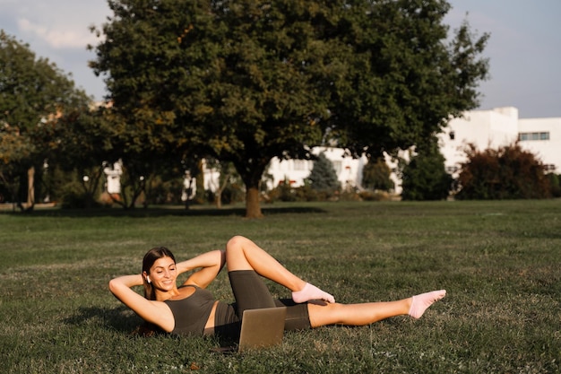 Online-Training mit Laptop im grünen Park Aktives fittes Mädchen macht Fitnesstraining im Freien