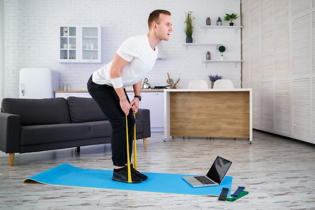 Onlinetraining. Junger Mann macht Übungen mit Fitness-Gummibändern mit Online-Tutorial zu Hause, freier Speicherplatz. Sport zu Hause machen