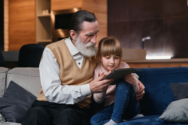 Opa mit seiner Enkelin mit Tablet-PC in gemütlichem Zuhause. Kleines Mädchen bringt ihrem Opa bei, intelligente Geräte zu benutzen.