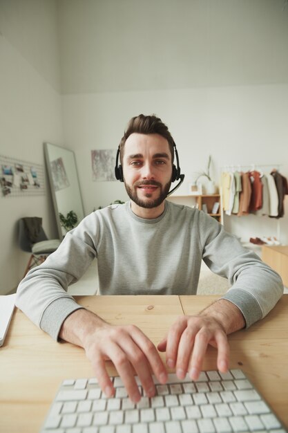 Operador amigable joven con auriculares escribiendo en el teclado de la computadora mientras está sentado en la mesa frente al monitor y consultando a los clientes en línea