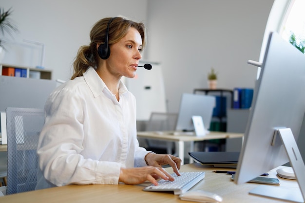 Operador de centro de llamadas femenino con auriculares de micrófono sentado en la computadora en la oficina