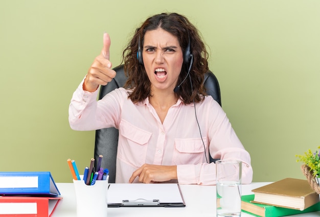 Foto operador de centro de llamadas femenino bastante caucásico molesto en auriculares sentado en el escritorio con herramientas de oficina apuntando al frente