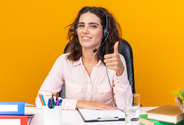 Operador de centro de llamadas femenino bastante caucásico sonriente en auriculares sentado en el escritorio con herramientas de oficina pulgar hacia arriba