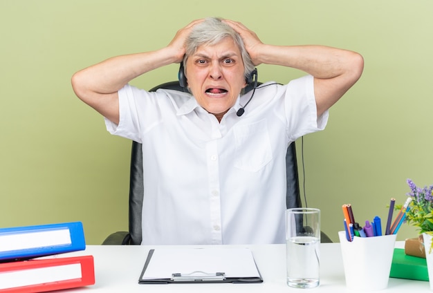Operador de centro de llamadas femenino caucásico ansioso en auriculares sentado en el escritorio con herramientas de oficina poniendo las manos sobre su cabeza