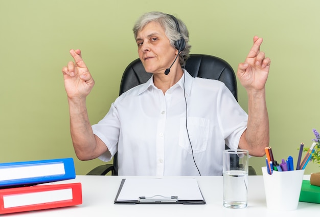 Operador de centro de llamadas femenino caucásico complacido en auriculares sentado en el escritorio con herramientas de oficina cruzando los dedos