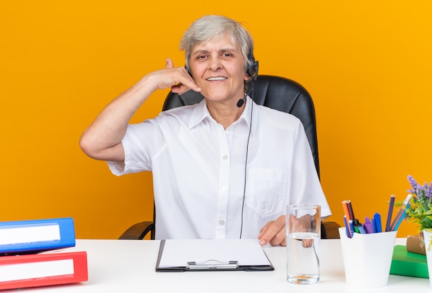 Operador de centro de llamadas femenino caucásico sonriente en auriculares sentado en el escritorio con herramientas de oficina gesticulando señal de llamada