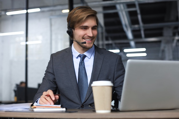 Operador de centro de llamadas masculino sonriente con auriculares sentado en la oficina moderna, consultar información en línea en una computadora portátil, buscar información en un archivo para ayudar al cliente.