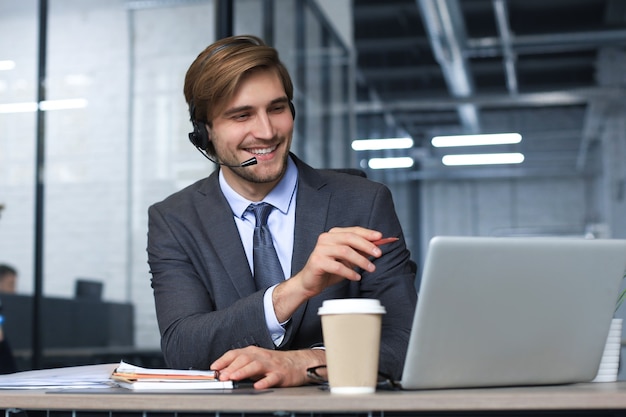 Operador de centro de llamadas masculino sonriente con auriculares sentado en la oficina moderna, consultar información en línea en una computadora portátil, buscar información en un archivo para ayudar al cliente.