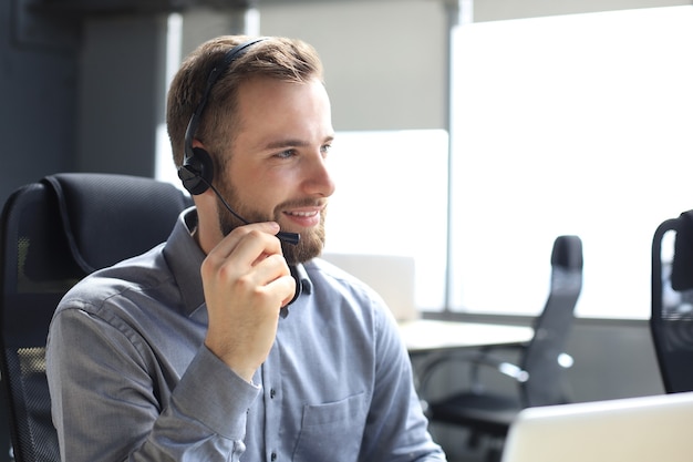 Operador de call center masculino sorridente com fones de ouvido sentado em um escritório moderno, consultando informações on-line em um laptop, procurando informações em um arquivo para ajudar o cliente