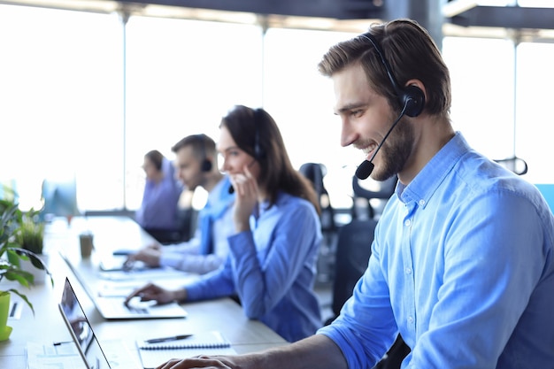 Operador de call center masculino sorridente com fones de ouvido sentado no escritório moderno com colegas no backgroung, consultoria online.