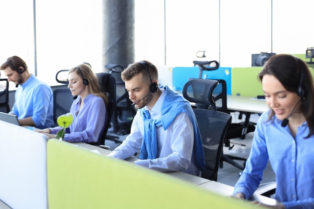 Operador de call center masculino sorridente com fones de ouvido sentado no escritório moderno com colegas no backgroung, consultoria online.