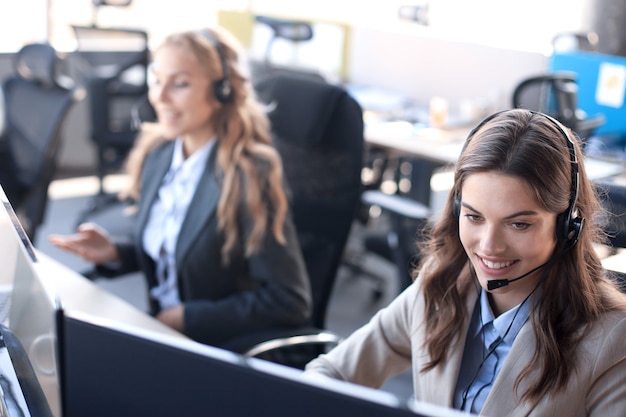 Operador de suporte ao cliente feminino com fone de ouvido e sorrindo, com colegas no fundo.