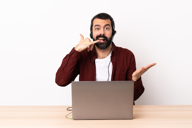 Operador de telemarketing homem caucasiano trabalhando com um fone de ouvido e com o laptop, fazendo gesto de telefone e duvidando