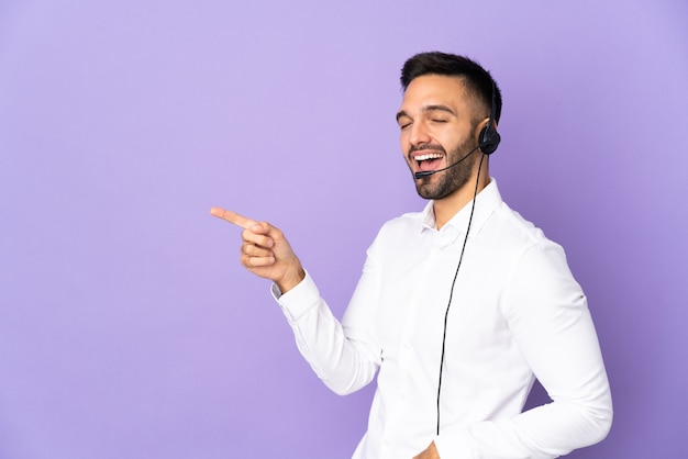 Operador de telemarketing trabalhando com um fone de ouvido isolado em um fundo roxo apontando o dedo para o lado e apresentando um produto