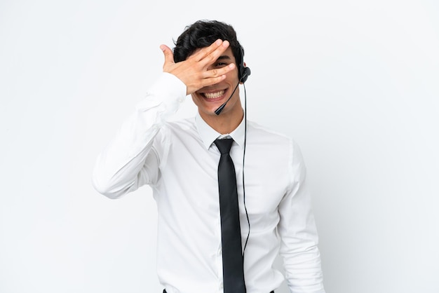 Operador de telemarketing trabalhando com um fone de ouvido isolado no fundo branco, cobrindo os olhos pelas mãos e sorrindo