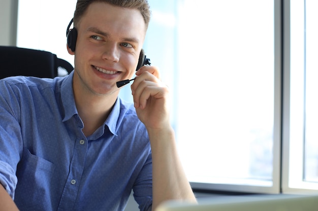 Operador masculino alegre del teléfono de la ayuda joven en el auricular, en el lugar de trabajo mientras usa la computadora portátil, el servicio de ayuda y el concepto del centro de llamadas de la consulta del cliente.