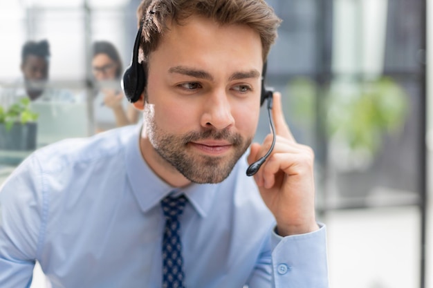 Operador masculino de telefone de suporte jovem alegre no fone de ouvido no local de trabalho com colegas em segundo plano enquanto usa o serviço de ajuda do computador e o conceito de call center de consultoria do cliente