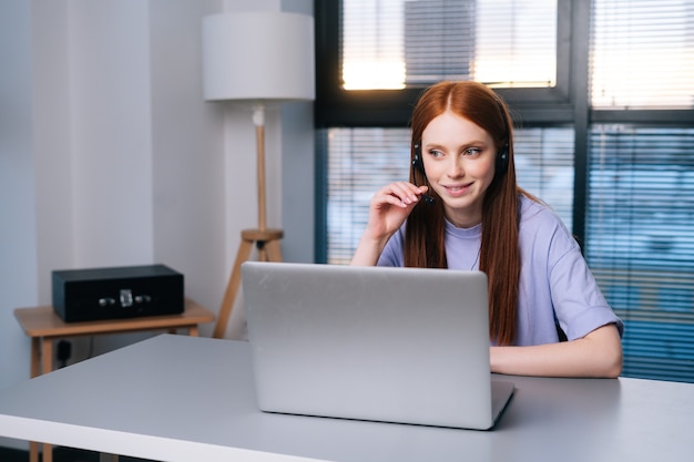 Operador de mujer joven atractiva con auriculares y computadora portátil durante la atención al cliente a través del teléfono. Representante de ventas en el servicio de asistencia hablando con micrófono y auriculares en el fondo de la ventana