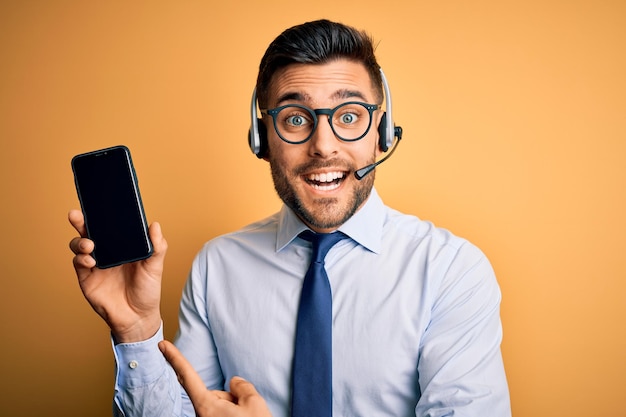 Operador de negocios hombre con auriculares de servicio al cliente del centro de llamadas que muestra la pantalla del teléfono inteligente muy feliz señalando con la mano y el dedo