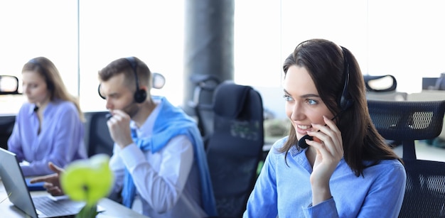 Operador de soporte al cliente femenino con auriculares y sonriendo, con colegas en segundo plano.