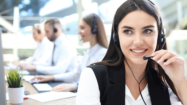 Operador de soporte al cliente femenino con auriculares y sonriendo, con colegas en segundo plano.