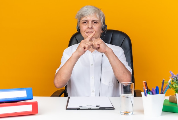 Operadora de call center feminina, caucasiana, satisfeita, com fones de ouvido, sentada na mesa, com as ferramentas de escritório, cruzando os dedos e gesticulando para não sinalizar