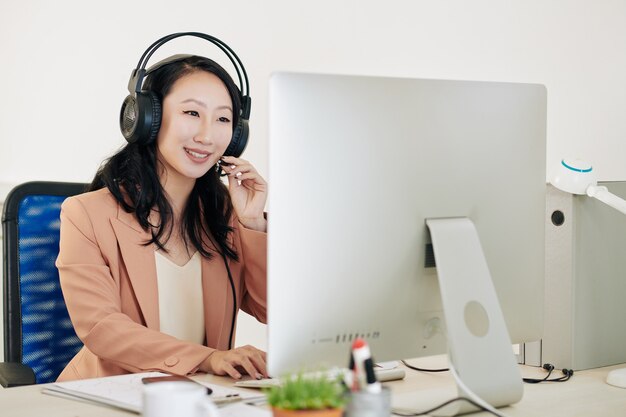 Operadora de suporte técnico asiática muito jovem e alegre em um fone de ouvido atendendo a chamada telefônica do cliente