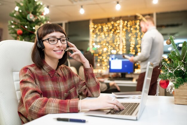 Operadora jovem feliz com fone de ouvido, consultando clientes on-line em frente ao laptop no escritório no dia de Natal