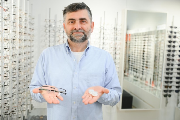 Ophthalmologisches Konzept Porträt eines Mannes, der zwischen Brillen und Kontaktlinsen wählt, der im Optikgeschäft steht und Kunststoffgehäuse und Spezifikationen in beiden Händen hält, die Brillen betrachten