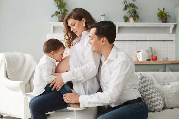 Opinião lateral a mulher gravida bonito que descansa com família em casa. menino beijando a barriga da mãe enquanto pai feliz sentado perto e abraçando a esposa. família alegre em camisas brancas. esperando bebê