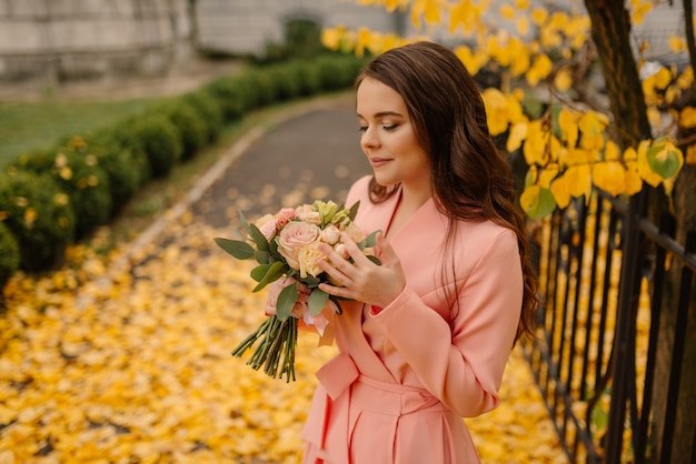 Opinión del retrato de una novia morena joven hermosa sensual en vestido de novia color de rosa y de pie en el parque de otoño cerca de la catedral vintage y con ramo de boda