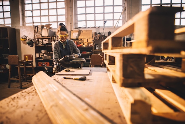 Opinión del retrato del trabajador de carpintería de sexo femenino profesional de mediana edad sonriente satisfecho con la prensa de acero en la tabla en el taller.