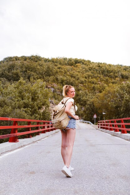 Opinión trasera la mujer rubia joven con la mochila que camina en el camino sobre un puente cerca de la montaña. Concepto de viaje y aventura. Viajero en medio del bosque. Viajar solo