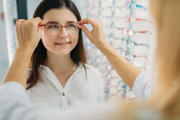 Optikerin und Kunde wählen Brillengestell gegen Schaufenster mit Brille im Optikgeschäft. Auswahl der Brille mit einem professionellen Optiker