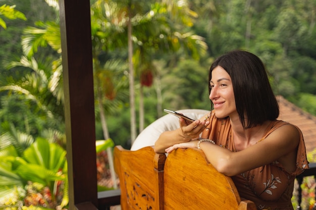 Optimismo. Atractiva mujer joven disfrutando del paisaje tropical y hablando por teléfono en su bungalow