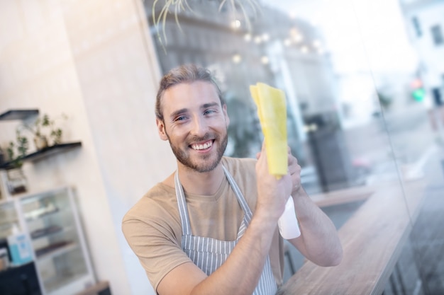 Optimista. Feliz joven adulto en camiseta y delantal de rayas limpiando el vidrio mientras está en el café