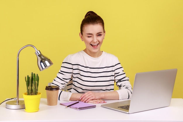 Optimistische Büroangestellte in gestreiftem Hemd, die am Arbeitsplatz sitzt und zwinkert