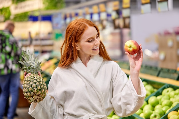 Optimistische Frau, die Früchte kauft, Ananas und Äpfel wählt, Bademantel trägt, lächelt