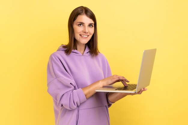 Optimistische junge erwachsene Frau, die an einem Laptop arbeitet und mit glücklichem Gesicht in die Kamera blickt