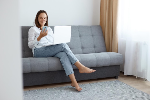 Optimistische, junge erwachsene Frau mit weißem Hemd und Jeans, die zu Hause auf dem Sofa sitzt und am Laptop arbeitet, eine Pause einlegt und einen Videoanruf auf dem Handy hat.