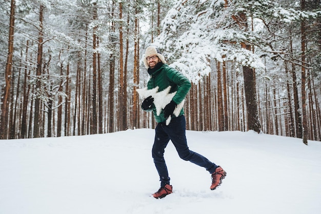 Optimistischer junger Mann in warmer Winterkleidung hat Spaß im Freien im Winterwald Atem frische Luft froh zu sehen, dass viel Schnee atrficial Tanne in Händen hält Menschen Saison Wetterkonzept