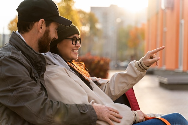 Optimistischer Mann, der seine schöne Frau umarmt, während sie während des Sitzens auf der Bank an der Straße interessiert die Straße hinunter zeigt