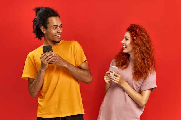 Optimistisches fröhliches Paar afroamerikanischer Mann und Ingwerfrau in einfachen T-Shirts, die sich gegenseitig anschauen, während sie Smartphones halten, chatten mit Anhängern aus dem Webblog. Technologie, Menschenkonzept