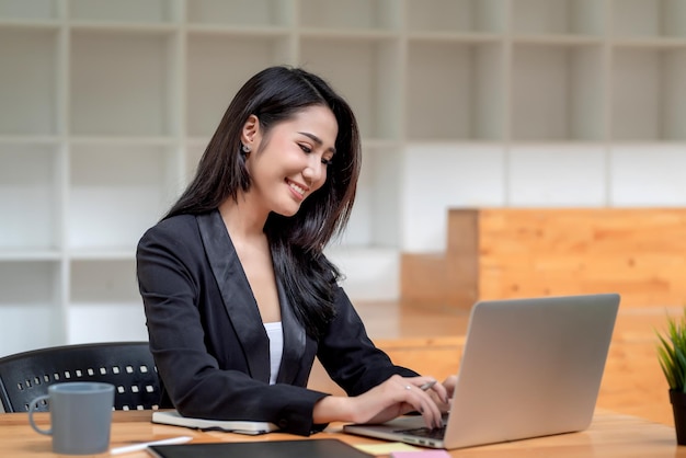 Optimistisches Sitzen der schönen jungen asiatischen Geschäftsfrau im Büro unter Verwendung einer Laptoptastatur.