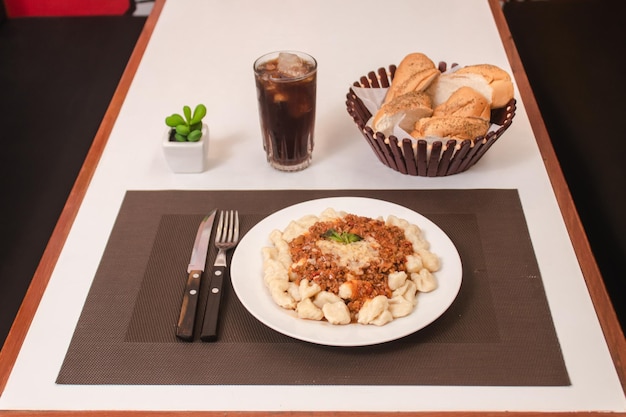 Ñoquis con salsa de carne con pan y soda sobre una mesa blanca