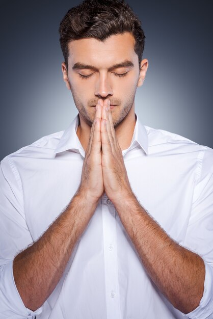 Foto orando por negocios. apuesto joven con camisa blanca cogidos de la mano cerca de la cara mientras está de pie contra el fondo gris