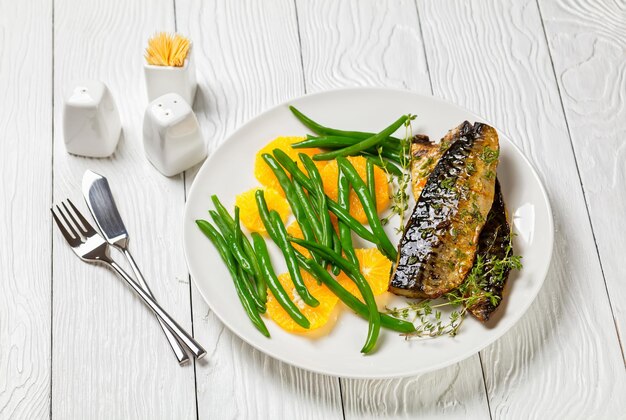 Orange glasiertes Makrelenfilet mit frischer Orange und gedämpften grünen Bohnen auf einem weißen Teller mit Besteck auf einem Tisch