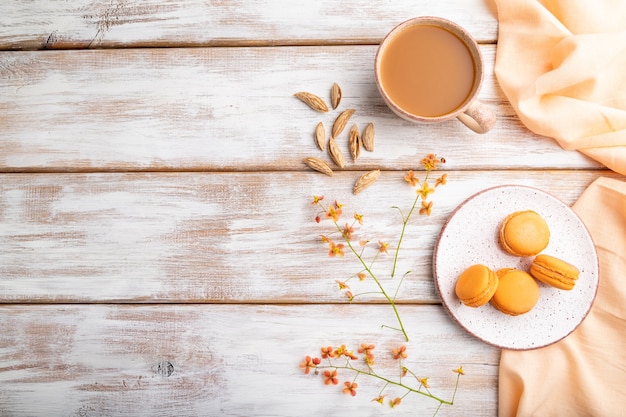 Orange Macarons oder Macaroons Kuchen mit Tasse Aprikosensaft auf einem weißen Holztisch und orange Leinen Textil. Draufsicht, flache Lage, Kopierraum.